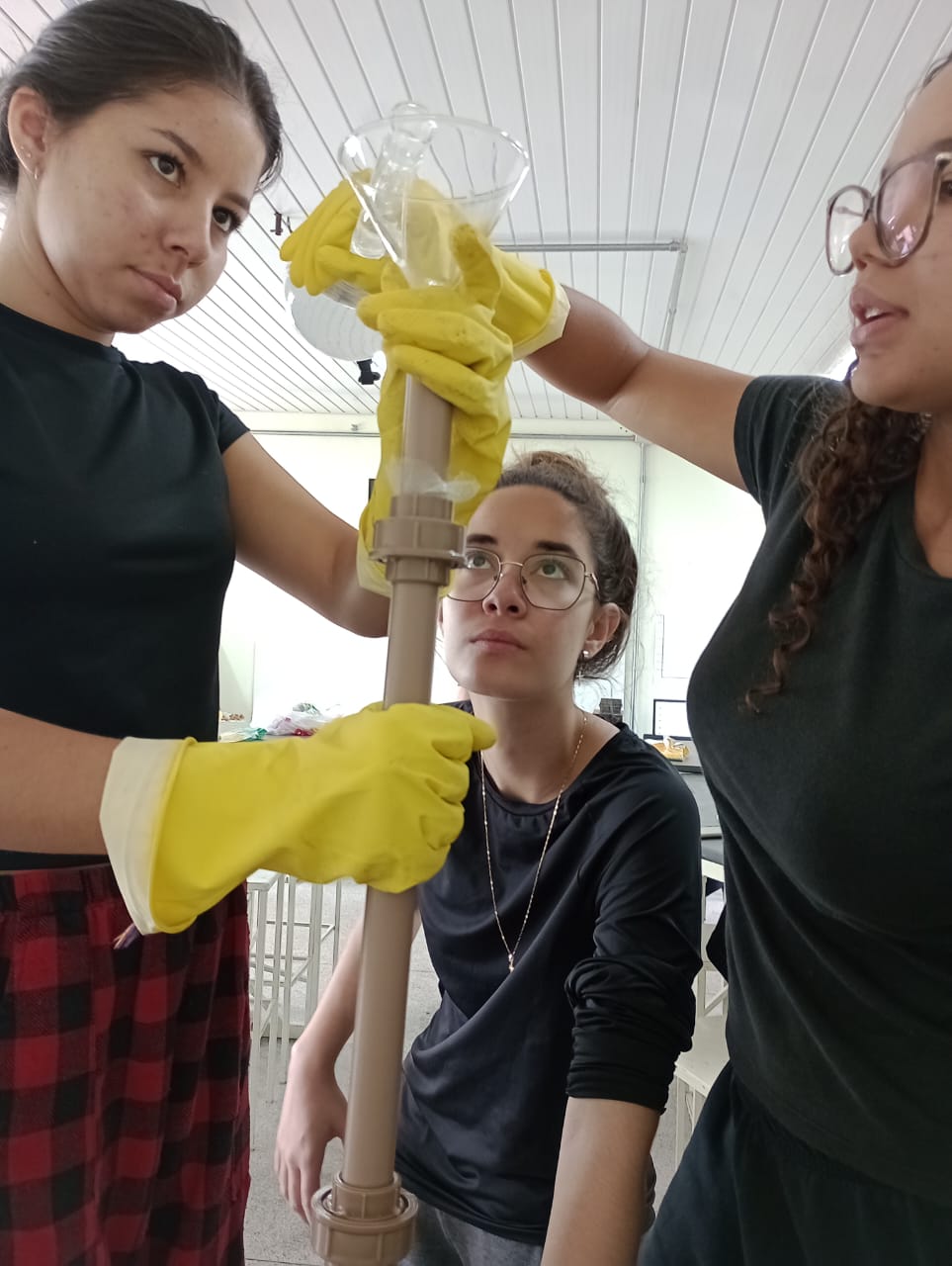 Três alunas testam o protótipo de filtro. Uma delas segura um béquer de vidro com água no topo do tubo de PVC, enquanto outra estabiliza o tubo. Todas usam luvas amarelas de proteção. A expressão delas indica concentração na execução do experimento.