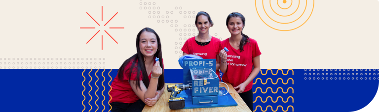 Tres jóvenes sonrientes se colocan alrededor de un proyecto tecnológico sobre una mesa de madera. Llevan camisetas rojas con el logotipo de Samsung y la frase Solve For Tomorrow impresa. En el centro, sobre la mesa, hay un dispositivo azul etiquetado con las palabras «PROPI-5» y «RE-FIVER» en letras blancas, con componentes como un tubo transparente y un taladro amarillo colocados a su lado. Los tres sostienen en la mano derecha pequeñas piezas de plástico azul, probablemente relacionadas con el proyecto. El fondo es decorativo, con formas geométricas sencillas en colores claros como el naranja y el azul.