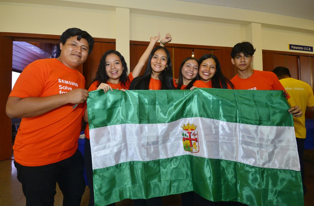 Seis estudiantes bolivianos con camisetas naranjas sostienen la bandera de Santa Cruz tras presentar su proyecto de tecnología asistiva en el Solve for Tomorrow Bolivia 2024.
