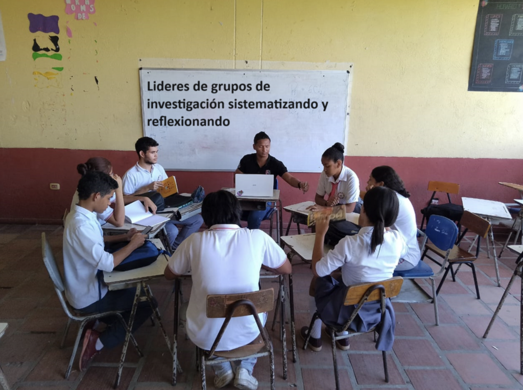 Grupo de estudiantes en un aula dialogando sobre temas relacionados con la lengua indígena, con cuadernos y carteles en las manos.